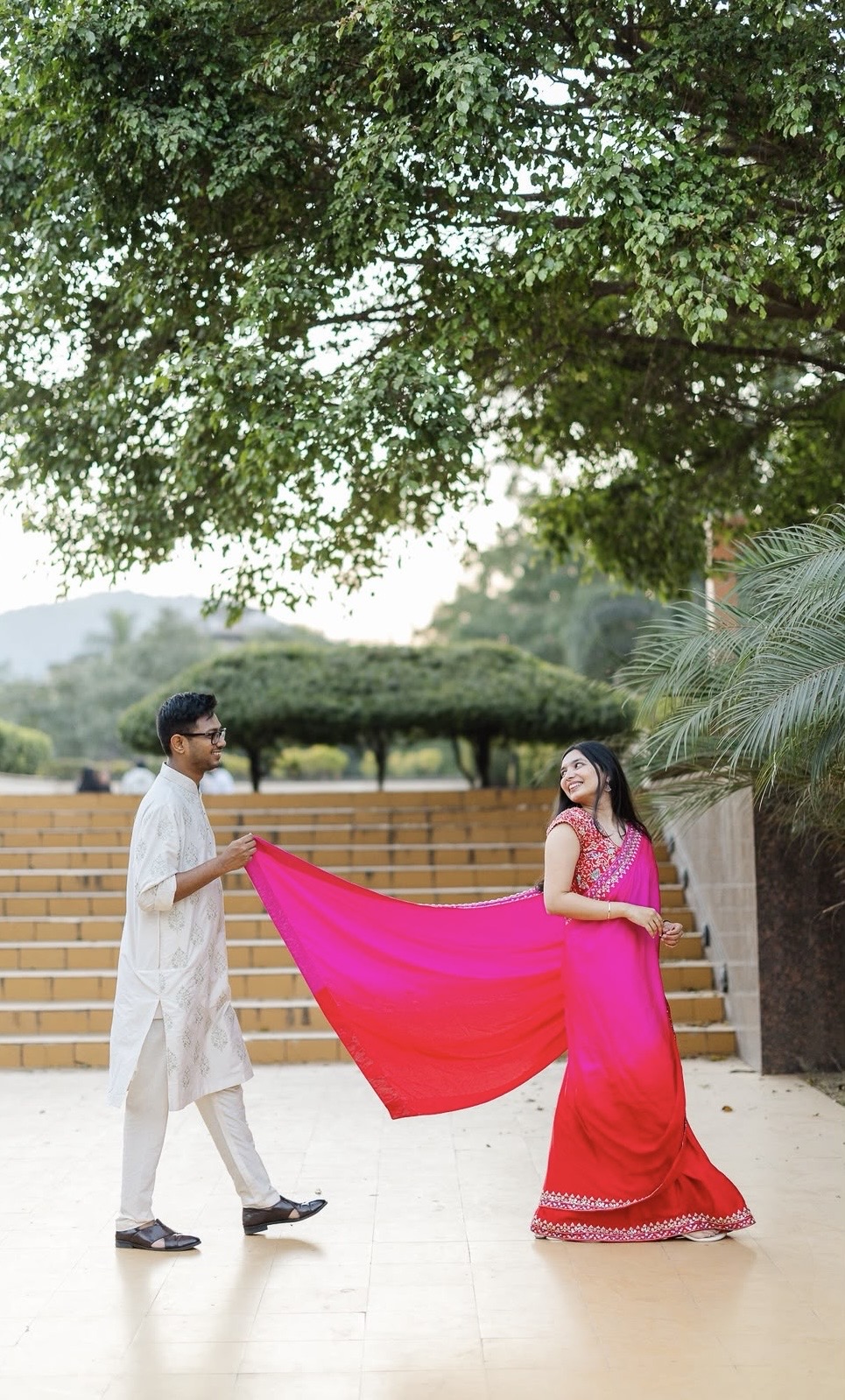 Saurabh holding Diksha’s bright pink sari as they smile at each other beneath the trees
